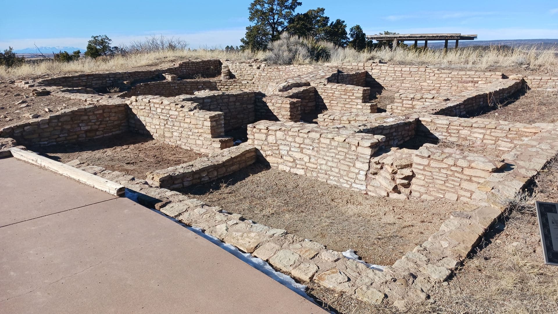 Excavated sandstone masonry room walls of Escalante Pueblo under a blue winter sky, with junipers, a shade ramada and distant snowy peaks behind