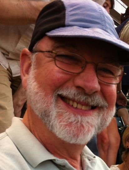 Bernard White Bell Jr. smiling outdoors, with a white beard, glasses and a blue and black cap