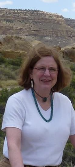 Carol Ambruster in Chaco Culture National Historic Park, smiling, in a white T-shirt and turquoise necklace, with canyon cliffs behind her