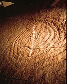 A spiral petroglyph pecked into sandstone, crossed by a vertical sliver of sunlight