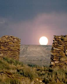 A full moon rising between two ruined stone masonry walls, with a mesa in the distance at dusk