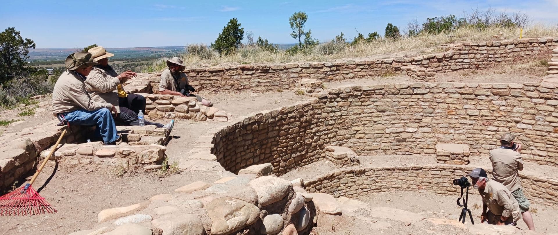 The SCAAS field crew at a circular stone-masonry kiva: three people talk on the wall while two photographers document it with a camera on a tripod
