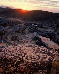Spiral petroglyphs on dark volcanic boulders with the sun setting over desert hills